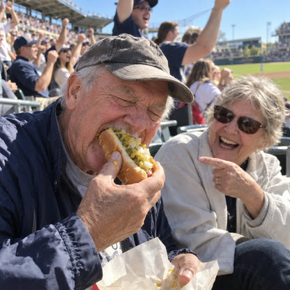 A man at a baseball game bites into a loaded hot dog while his wife laughs beside him